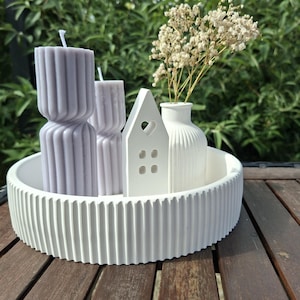 Könnte beinhalten: Ein dekoratives weißes Tablett präsentiert eine Sammlung von Gegenständen. Es gibt zwei lavendelfarbene Säulenkerzen, ein weißes Keramikhaus mit herzförmigen Ausschnitten und eine kleine weiße Vase mit Trockenblumen. Das Tablett hat ein geripptes Design.