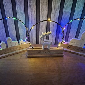 Könnte beinhalten: Drei dekorative Lichtdisplays aus Holz. Jedes hat einen Holzsockel mit einem weißen Ausschnittdesign, darunter eine Kirche, Häuser und ein Rentier. Ein kreisförmiger Drahtrahmen mit farbigen LED-Leuchten umgibt jedes Design.