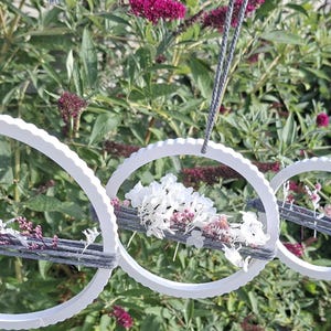 Könnte beinhalten: Eine Reihe weißer, kreisförmiger Hängedekorationen mit strukturiertem Rand. Jeder Ring hat einen grauen, gewebten Abschnitt, der mit weißen und lila Blumen verziert ist und an grauen Kordeln aufgehängt ist. Der Hintergrund ist ein üppiger grüner Garten.