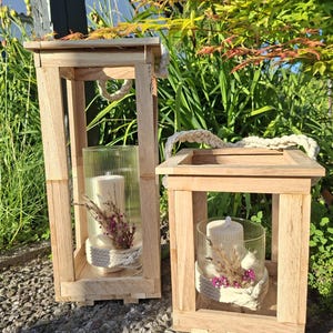 Pode incluir: Duas lanternas de madeira com castiçais de vidro. Cada lanterna tem uma alça de corda grossa e contém uma vela branca, flores secas e uma base de corda decorativa. As lanternas estão sobre uma superfície de pedra com vegetação ao fundo.