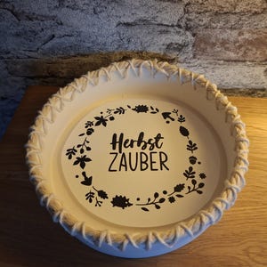 Pode incluir: Uma bandeja decorativa redonda, creme, com uma borda de corda trançada. O centro da bandeja apresenta as palavras "Herbst ZAUBER" em preto, rodeadas por um desenho floral e de folhas. A bandeja está sobre uma superfície de madeira.