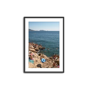 May include: Framed photograph of a coastal scene with people on rocky cliffs and swimming in the turquoise water. A blue inflatable ring rests on the rocks. A mountain range is visible in the distance under a clear blue sky.