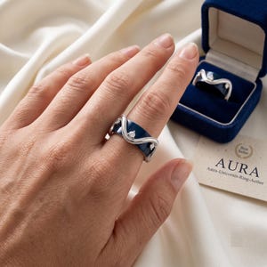May include: A wide band ring featuring a deep blue enamel inlay with a silver wavy border and three small clear stones. The ring is displayed on a finger and inside a blue velvet box with a card labeled AURA Astra-Unicornis-Ring-Aether.
