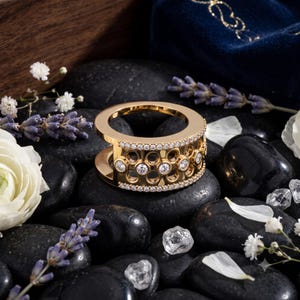 May include: A gold-toned ring featuring a wide, open-work band with a honeycomb-inspired pattern. The ring is accented with two rows of small, clear, round-cut stones along the edges and a central row of larger, round-cut stones set within the band.