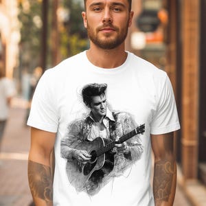May include: A white short-sleeved t-shirt featuring a black and white sketch-style graphic of a musician with a pompadour hairstyle playing an acoustic guitar. The design is centered on the chest, showcasing detailed shading and artistic line work.