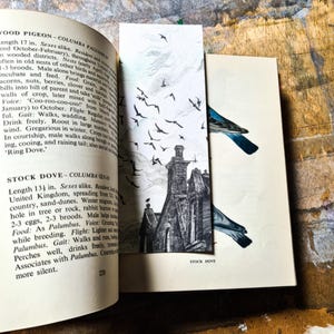 May include: A vintage bookmark featuring a black and white illustration of a building with birds flying above it. A second bookmark shows a blue bird. The bookmarks are placed inside an open book with text.