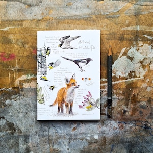 May include: A white notepad featuring the words "Urban Wildlife" and drawings of birds, a fox, and a butterfly. A dark grey pencil lies on a wooden surface beside the notepad.
