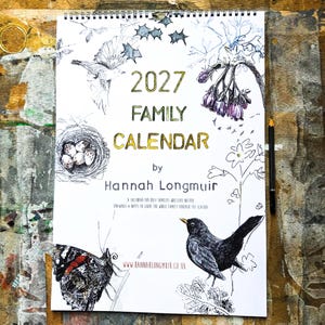 May include: A 2027 family calendar by Hannah Longmuir with a spiral binding. The cover features black ink illustrations of a bird, a butterfly, a bird's nest with eggs, and purple flowers. The text reads 2027 FAMILY CALENDAR by Hannah Longmuir.