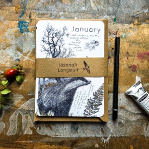 May include: A set of illustrated cards featuring a badger and a tree, held by a brown paper band labelled Hannah Longmuir with a bird icon. The top card reads January and includes handwritten text. A black pencil and a paint tube are placed nearby.