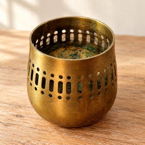 May include: A cylindrical brass candle holder with a weathered, antique gold finish. The upper section features a repeating pattern of vertical cutouts and small circular perforations, creating a decorative band around the top rim of the vessel.