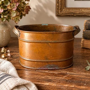 May include: An oval-shaped copper basin featuring two side handles and a weathered, metallic finish. The vessel displays visible signs of oxidation with subtle green patina accents along the base and rim, showcasing a rustic, aged appearance.