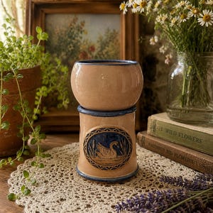 May include: A two-piece ceramic wax warmer with a light tan glaze and dark blue trim. The cylindrical base features a circular relief medallion depicting a swan swimming in a pond with reeds. The top section is a bowl for wax melts.