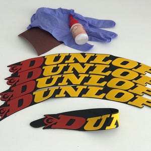 May include: A collection of Dunlop motorcycle tire decals in red, yellow, and black. The image also includes a pair of blue gloves, a small bottle of adhesive, and a piece of sandpaper, suggesting a repair or customization kit.