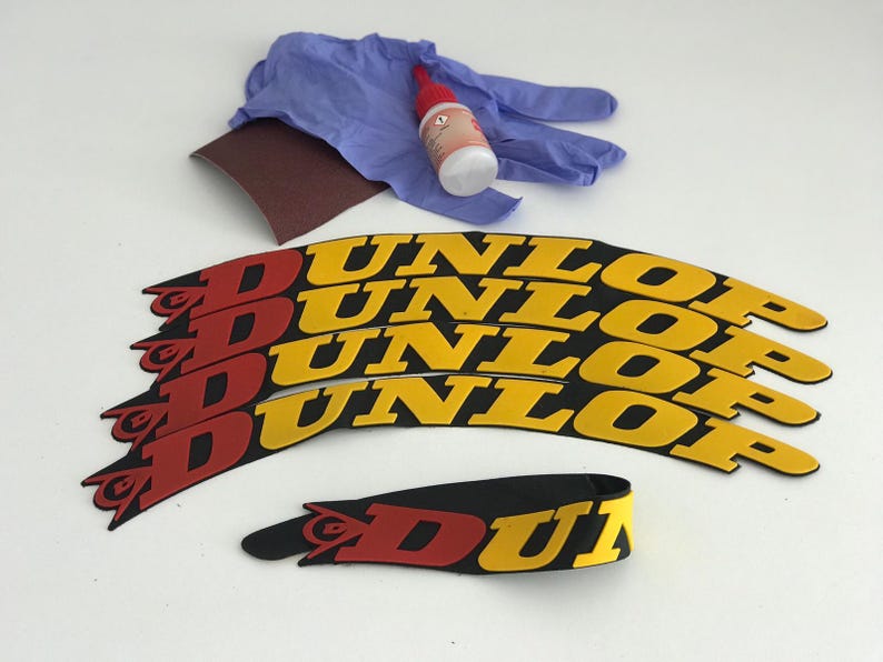 May include: A collection of Dunlop motorcycle tire decals in red, yellow, and black. The image also includes a pair of blue gloves, a small bottle of adhesive, and a piece of sandpaper, suggesting a repair or customization kit.