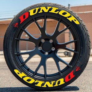 May include: A black car wheel with a Dunlop tire. The tire has yellow and red Dunlop lettering. The wheel has a black, multi-spoke design. The tire is on a paved surface.