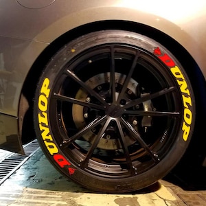May include: Close-up of a black car wheel with a Dunlop tire. The tire has yellow and red lettering that reads "DUNLOP". The wheel is black with multiple spokes. The car's body is gray.
