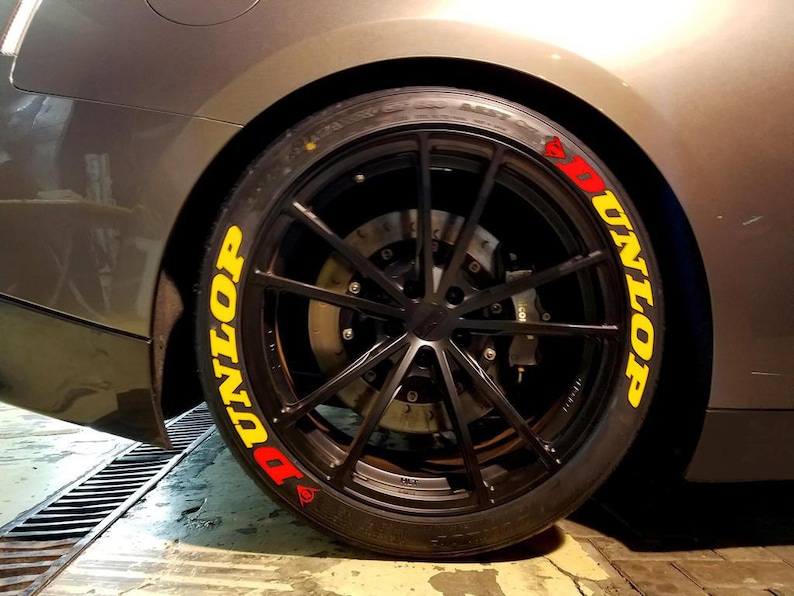 May include: Close-up of a black car wheel with a Dunlop tire. The tire has yellow and red lettering that reads "DUNLOP". The wheel is black with multiple spokes. The car's body is gray.