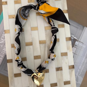 Könnte beinhalten: Ein gemustertes Tuch in Schwarz, Weiß und Gelb mit einem goldenen Herzanhänger. Das Tuch ist oben geknotet und bildet eine Halskette. Der Anhänger ist ein großes, glänzendes Herz.