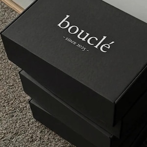 Puede incluir: Una pila de tres cajas rectangulares de cartón negro. La caja superior presenta el texto blanco "bouclé" en una fuente serif, con el texto "- since 2025 -" impreso directamente debajo en una fuente serif blanca a juego.