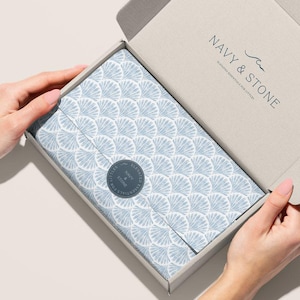 Op de afbeelding: Een lichtgrijze kartonnen doos met een open deksel, met een pakket verpakt in lichtblauw en wit patroonpapier. Het papier heeft een waaierachtig ontwerp. De doos heeft de tekst "NAVY & STONE" op het deksel.