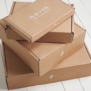May include: A stack of three rectangular brown cardboard shipping boxes. The top box features the white text 'nu-in' and 'Fashion first. Sustainable always.' Each box is decorated with a small white leaf icon printed on the front edge.