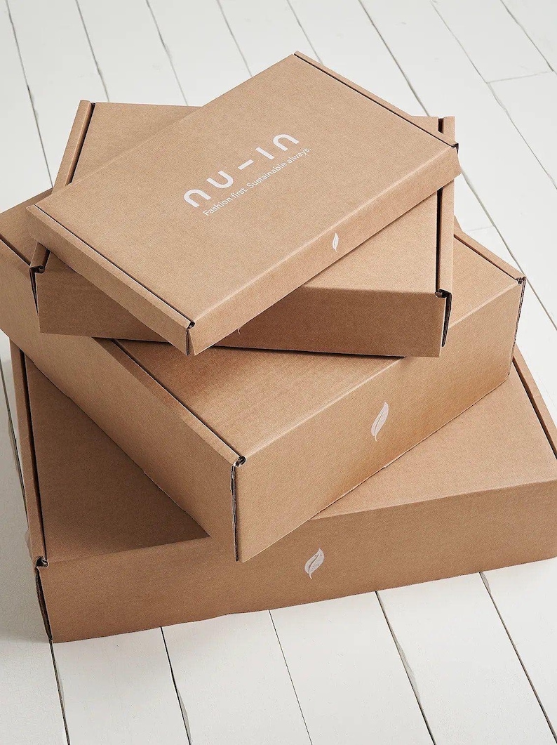 May include: A stack of three rectangular brown cardboard shipping boxes. The top box features the white text 'nu-in' and 'Fashion first. Sustainable always.' Each box is decorated with a small white leaf icon printed on the front edge.