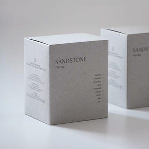 Puede incluir: Dos cajas grises SANDSTONE Drip Bag se muestran sobre una superficie blanca. Las cajas tienen el nombre de la marca y la descripción del producto en una fuente minimalista. Las cajas enumeran los orígenes del café, incluyendo Panamá, Colombia y Costa Rica.
