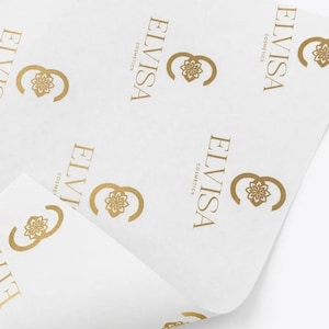May include: A sheet of white tissue paper featuring a repeating pattern of the gold-colored logo for ELVISA COSMETICS. The logo consists of a stylized floral emblem next to the text ELVISA COSMETICS printed in a clean, sans-serif font.