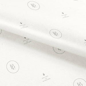 May include: White tissue paper featuring a repeating pattern of black logos. Each logo consists of a circular emblem containing the letters BL, accompanied by the text Enliven Your Interiors and a small floral icon printed in black ink.