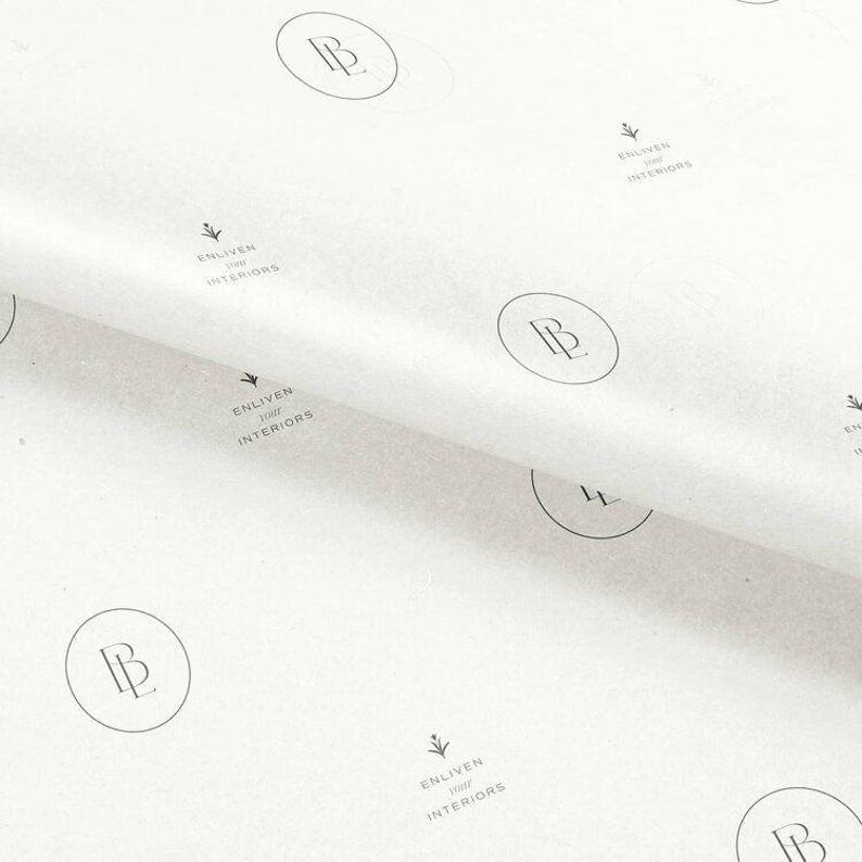 May include: White tissue paper featuring a repeating pattern of black logos. Each logo consists of a circular emblem containing the letters BL, accompanied by the text Enliven Your Interiors and a small floral icon printed in black ink.