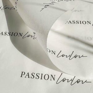 May include: A sheet of white tissue paper with a repeating pattern of the text PASSION London printed in a combination of bold sans-serif and elegant cursive script. The paper is partially rolled, creating a curved fold across the center.