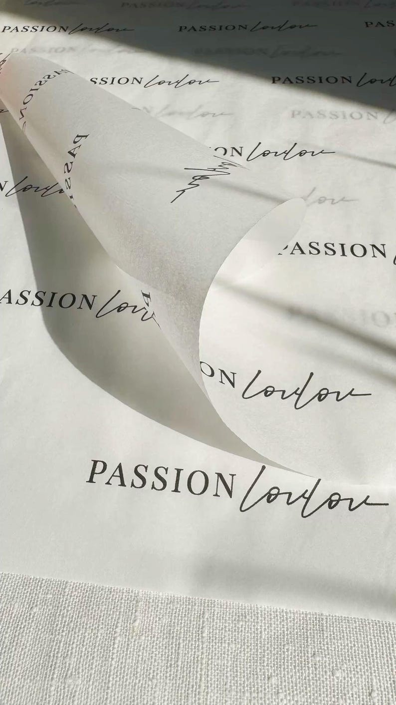 May include: A sheet of white tissue paper with a repeating pattern of the text PASSION London printed in a combination of bold sans-serif and elegant cursive script. The paper is partially rolled, creating a curved fold across the center.