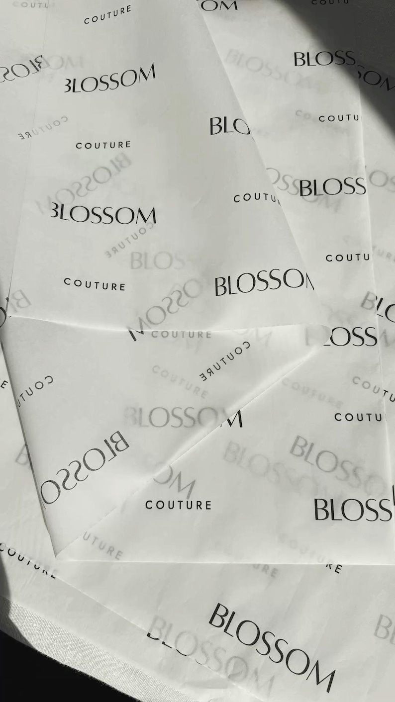 May include: White tissue paper featuring a repeating black print of the word Blossom followed by the word Couture. The text appears in various orientations, including some instances that are mirrored or partially obscured by folds in the paper.