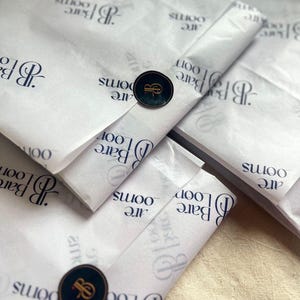 May include: Multiple rectangular packages wrapped in white tissue paper featuring a repeating dark blue logo and the text Bare Looms. Each package is sealed with a circular black sticker displaying a gold letter B logo with a small red accent.