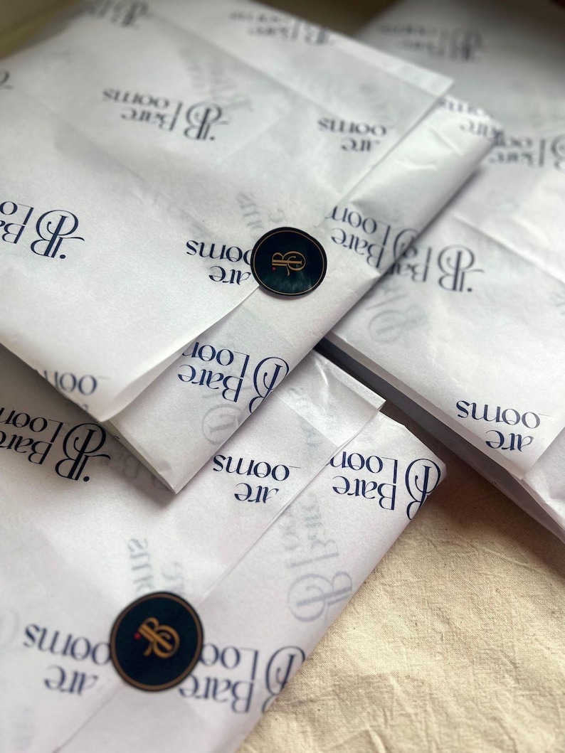 May include: Multiple rectangular packages wrapped in white tissue paper featuring a repeating dark blue logo and the text Bare Looms. Each package is sealed with a circular black sticker displaying a gold letter B logo with a small red accent.