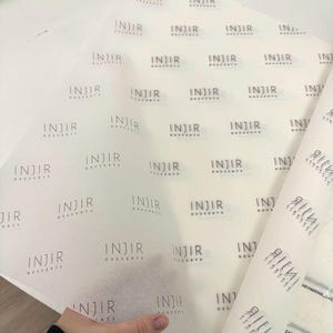 May include: A sheet of translucent white tissue paper featuring a repeating black logo that reads INJIR DESSERTS. The paper is thin and semi-transparent, showing the brand name printed in a clean, sans-serif font across the entire surface.