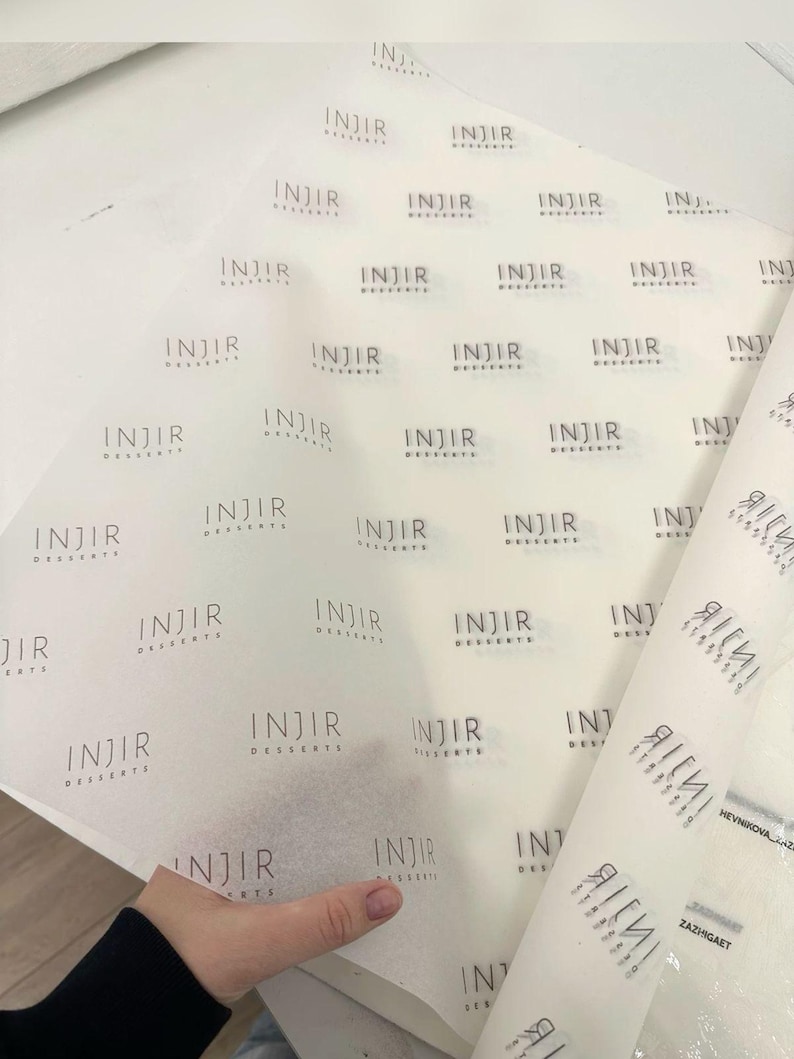 May include: A sheet of translucent white tissue paper featuring a repeating black logo that reads INJIR DESSERTS. The paper is thin and semi-transparent, showing the brand name printed in a clean, sans-serif font across the entire surface.