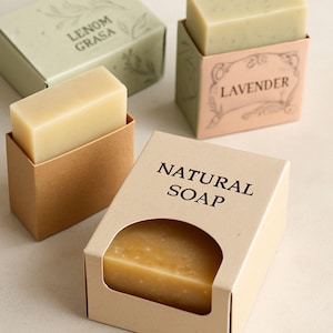 May include: Three rectangular bars of soap in individual paper packaging. One bar is in a tan box labeled NATURAL SOAP, another in a brown box with a cream bar, and the third in a light green box labeled LENOM GRASA, plus a lavender labeled box.