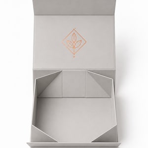 Peut inclure: Une boîte cadeau carrée gris clair ouverte avec un couvercle à charnière. L'intérieur du couvercle présente un logo en ligne de couleur cuivre métallique représentant trois feuilles dans un losange, avec de petites étoiles en haut et en bas.
