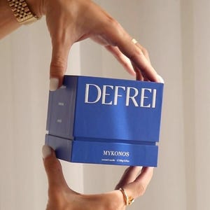 Puede incluir: Una caja cuadrada azul para vela aromática con el texto blanco DEFREI impreso en el frente. Debajo del nombre de la marca, el texto dice MYKONOS y scented candle 150g 5.3oz. La caja tiene un diseño de dos piezas con tapa y base.