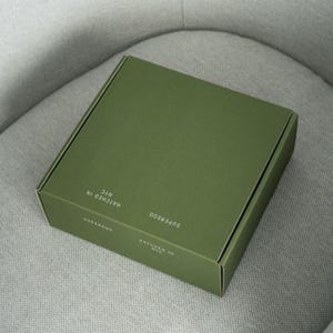 May include: A square, olive green cardboard shipping box with a matte finish. The top and side panels feature white text that reads SUPEREGG and HATCHED IN NYC. The box is closed and has a clean, structured design with sharp, folded edges.