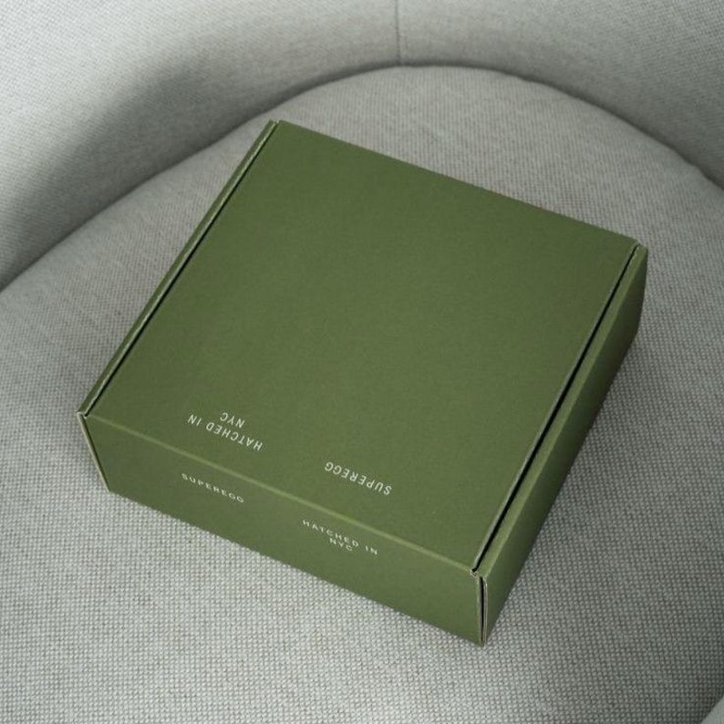 May include: A square, olive green cardboard shipping box with a matte finish. The top and side panels feature white text that reads SUPEREGG and HATCHED IN NYC. The box is closed and has a clean, structured design with sharp, folded edges.