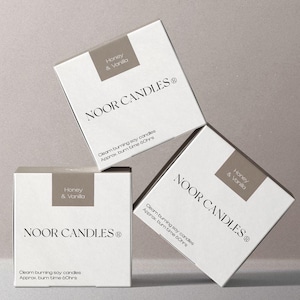 May include: Three white square boxes of Noor Candles with a grey label reading Honey & Vanilla. The text reads Noor Candles, clean burning soy candles, and approx. burn time 60hrs. The boxes are arranged in a stacked and angled composition.