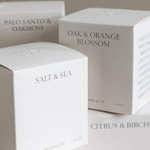 May include: Close-up of four white square boxes with black text. The boxes are labeled "PALO SANTO & OAKMOSS", "OAK & ORANGE BLOSSOM", "SALT & SEA", and "CITRUS & BIRCH". Each box has the McGee & Co. logo and the weight of the candle, 8 oz (227 g).