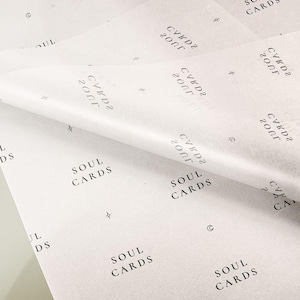 May include: A sheet of white tissue paper featuring a repeating black print of the text SOUL CARDS, along with small star and crescent moon symbols. The paper is folded over, showing a semi-transparent texture and a consistent pattern throughout.