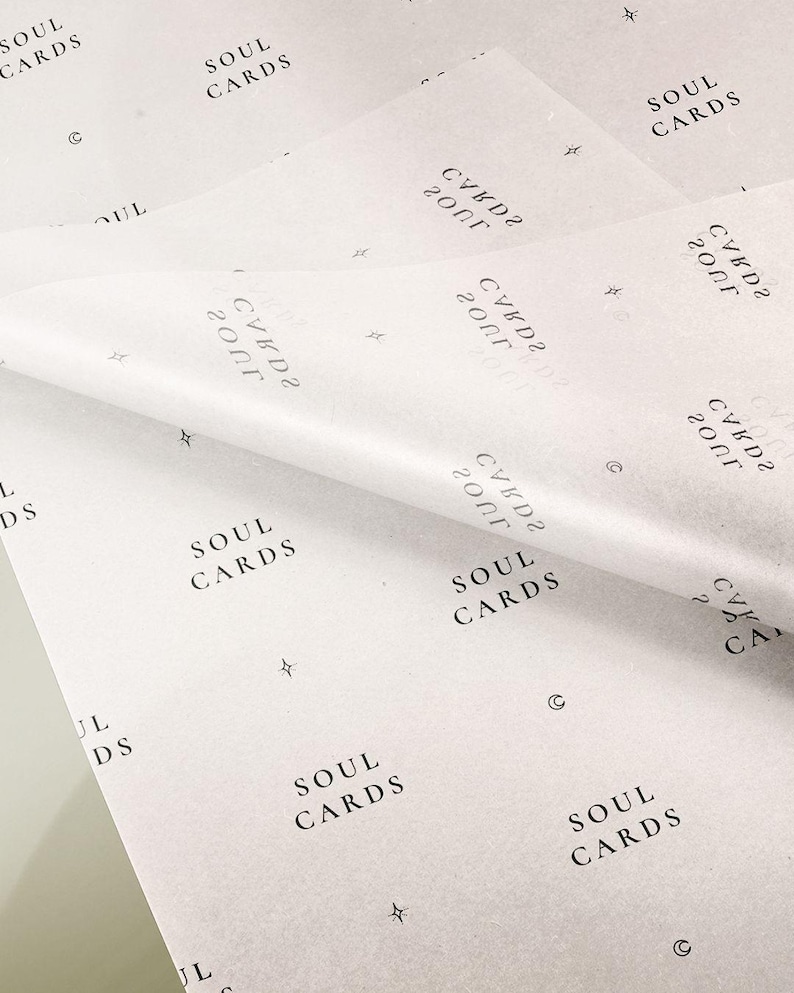 May include: A sheet of white tissue paper featuring a repeating black print of the text SOUL CARDS, along with small star and crescent moon symbols. The paper is folded over, showing a semi-transparent texture and a consistent pattern throughout.