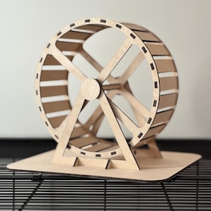 May include: A wooden hamster wheel with a natural finish. The wheel has a circular design with horizontal slats and is supported by a base. The wheel is designed for small pets and is placed on a black wire surface.