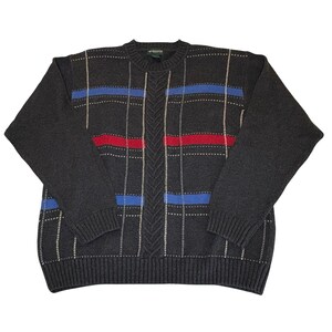 May include: A charcoal gray long-sleeve crew neck sweater featuring a vertical cable knit pattern down the center, accented by horizontal stripes in bright blue and red, and thin vertical white lines forming a subtle plaid-like grid pattern.