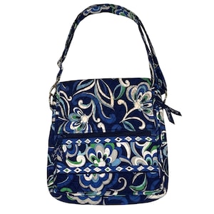 May include: A navy blue crossbody bag with a floral pattern in shades of blue, green, and white. The bag has a long adjustable strap, a front zippered pocket, and a flap closure. The bag is made of quilted fabric.