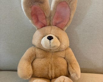 May include: A plush stuffed rabbit toy with tan fur, long upright ears featuring pink inner lining, a rounded snout with a dark nose, and pink accents on the bottom of its feet. The toy is seated in an upright position with a soft, fuzzy texture.